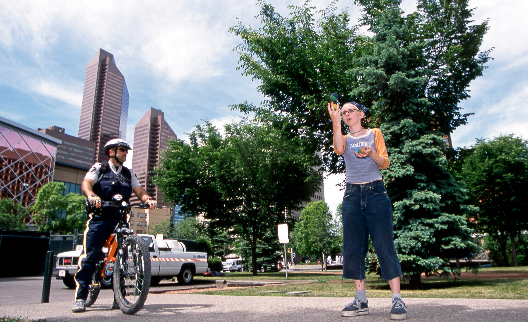 A police officer on a bicycle stands to the left of the frame while a person in casual streetwear juggles to the right.