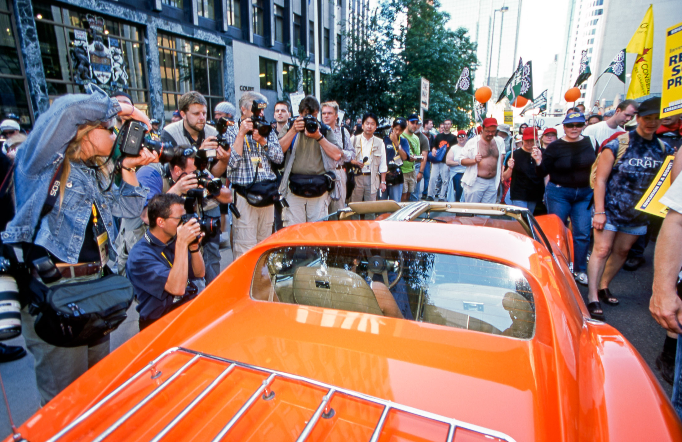 An orange sports car is surrounded by photojournalists and protesters on a street in downtown Calgary.