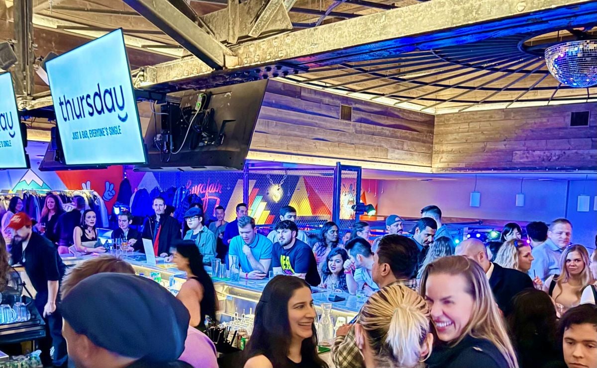A crowded bar is filled with people standing and chatting with each other. Two white digital screens over the bar read “Thursday: Just a bar, everyone’s single.”