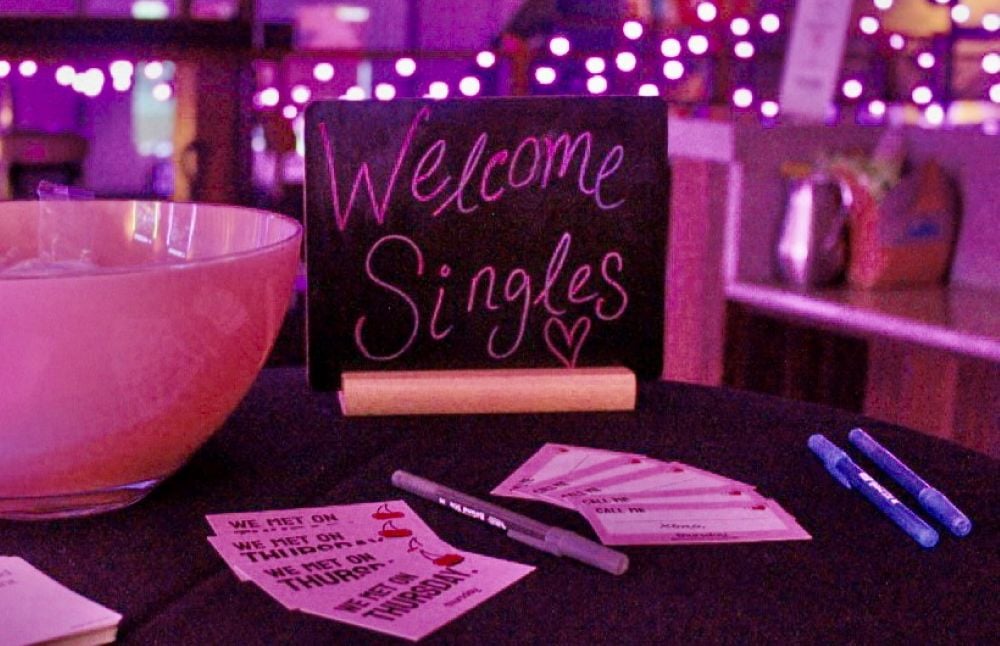 A small black chalkboard sits on a table in a dim room with pink lighting amidst a bowl of mints, business cards and pens. The blackboard has “Welcome singles” written on it. In the background are strings of lights.