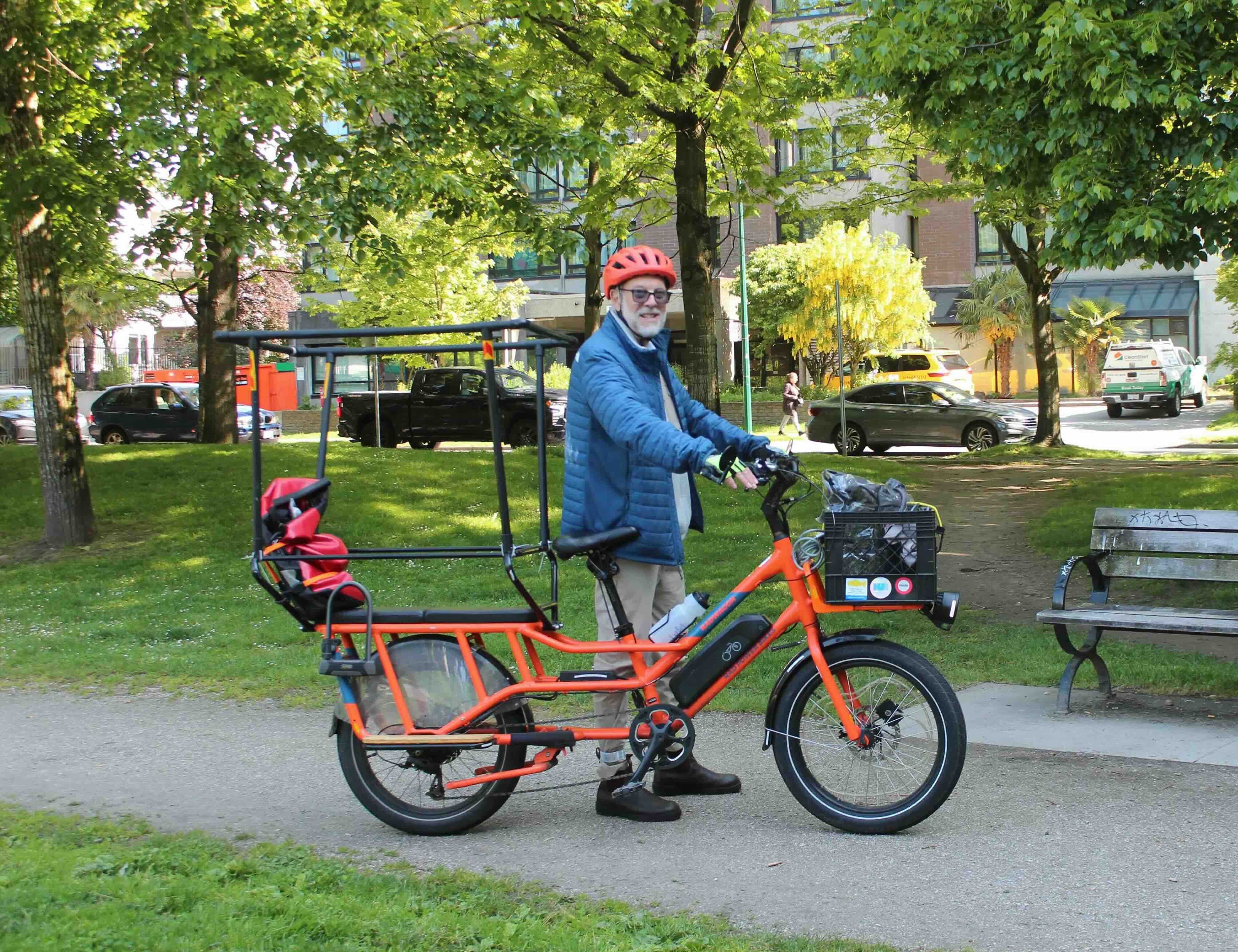 David Finnis stands with his electric cargo bike, an orange bicycle with bench seating at the back. He is wearing a red helmet and a blue puffer jacket.