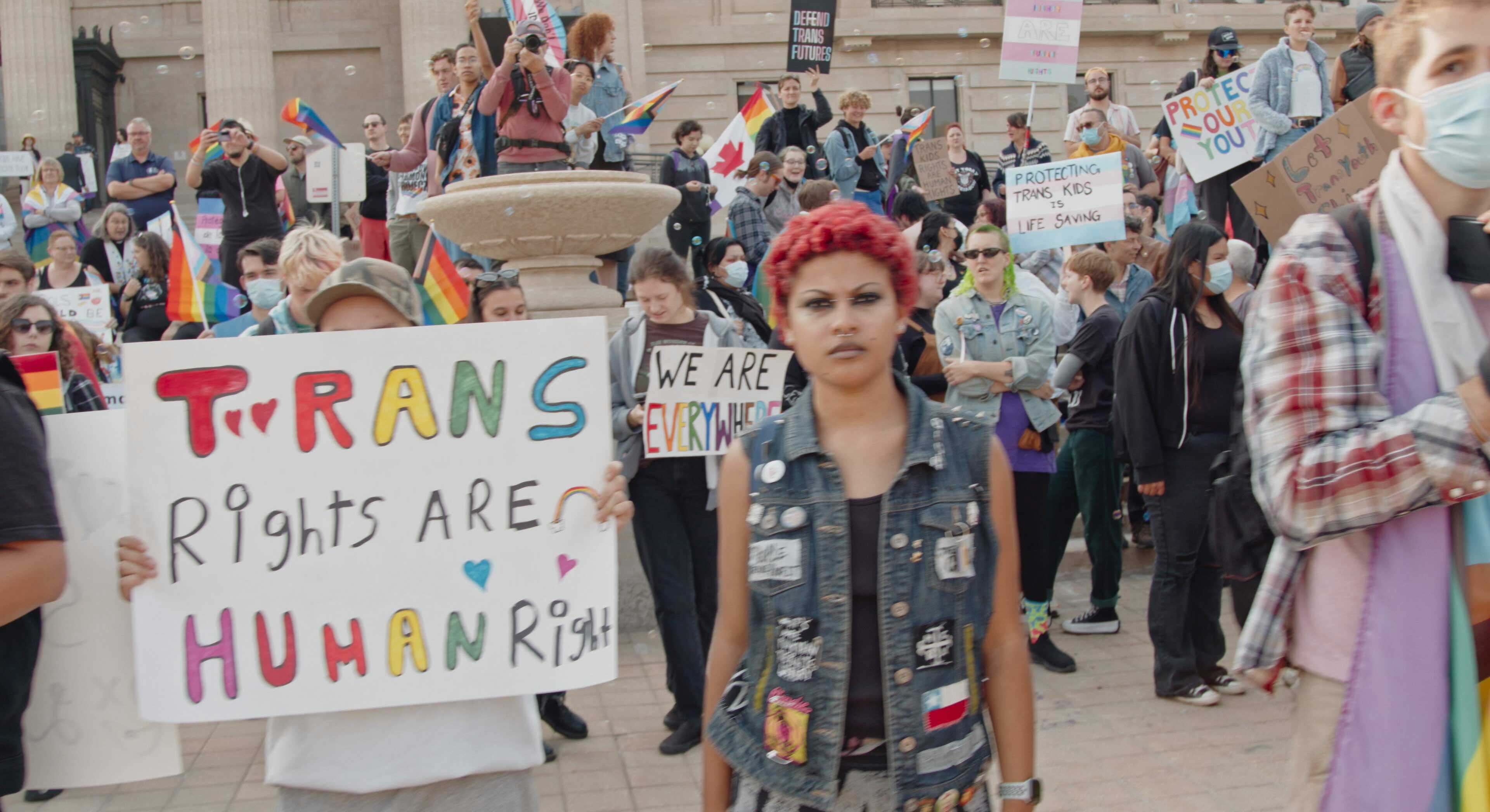 Crowds of people in casual streetwear crowd a public outdoor space holding white protest signs that read “Trans rights are human rights,” “We are everywhere,” “Protecting trans kids is lifesaving,” and more. Some are holding rainbow flags or flags with pink, white and light blue stripes. 
