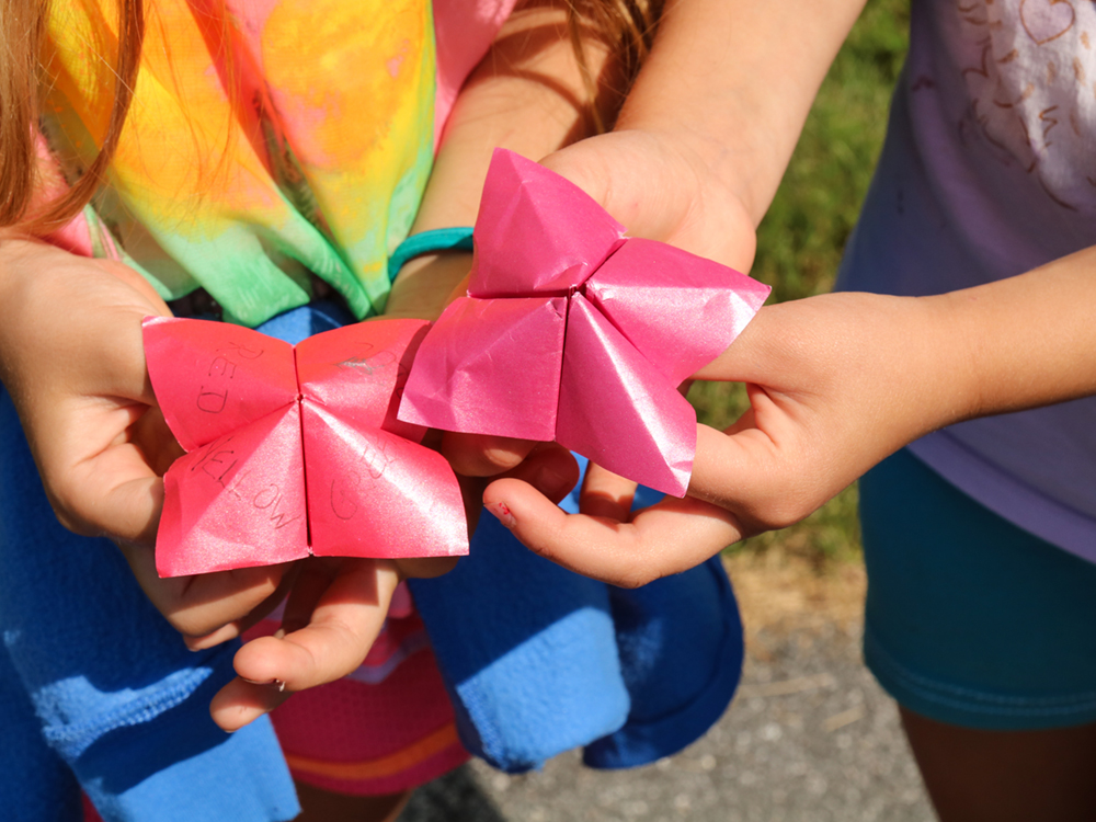 The hands of two children hold up red paper &ldquo;fortune tellers,&rdquo; paper crafts with four folded quadrants. They are wearing colourful summer clothes on a bright day.