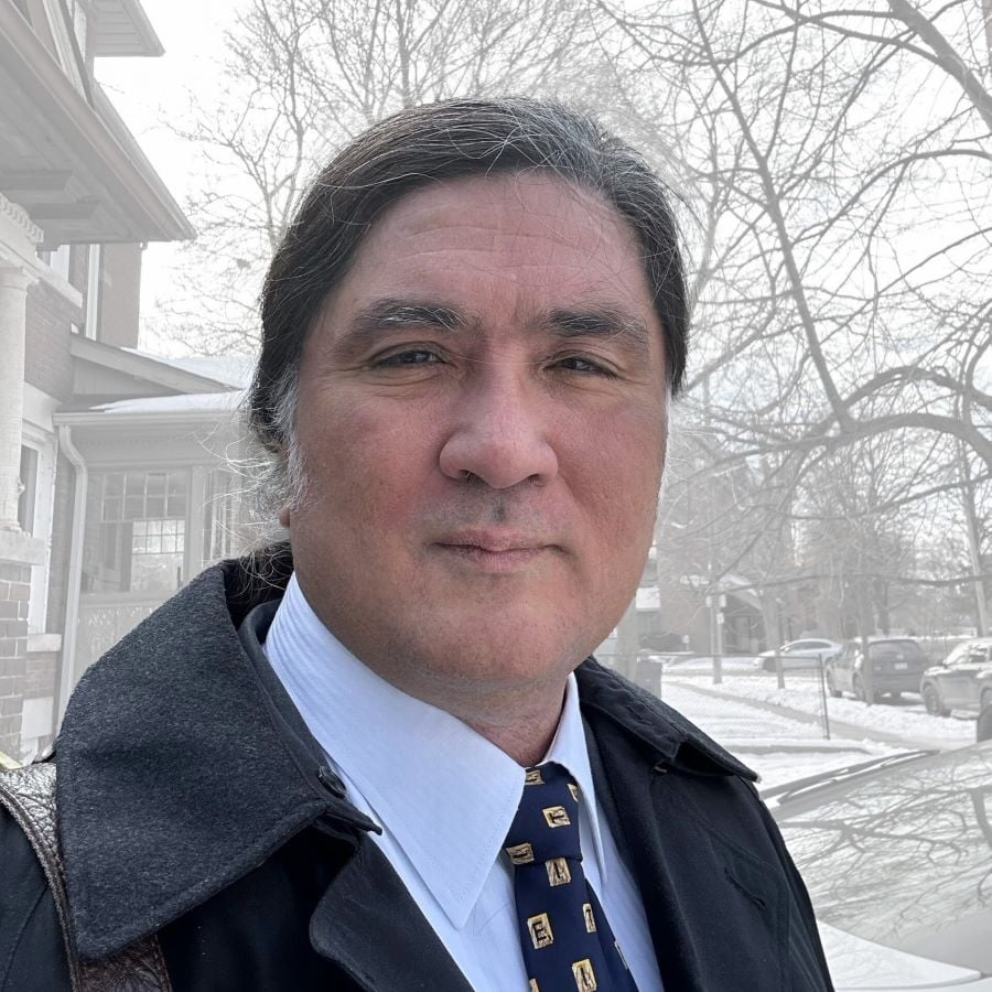 A man with medium-light skin tone and dark hair, wearing a dark grey coat over a shirt and tie, looks directly into the camera, with a snowy residential neighbourhood in the background.