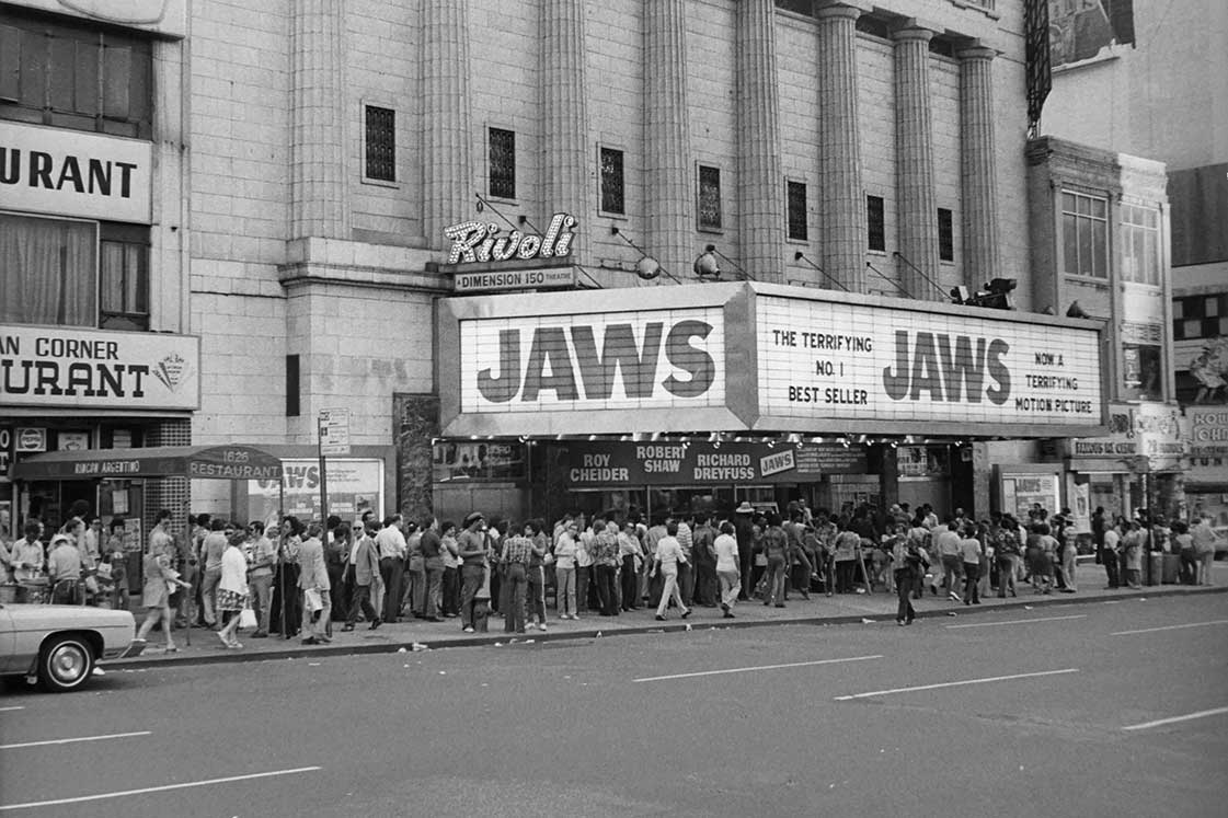 A black-and-white photo of crowds of people lined up around the block for a screening of Jaws. The movie’s title is large across a movie theatre marquee.