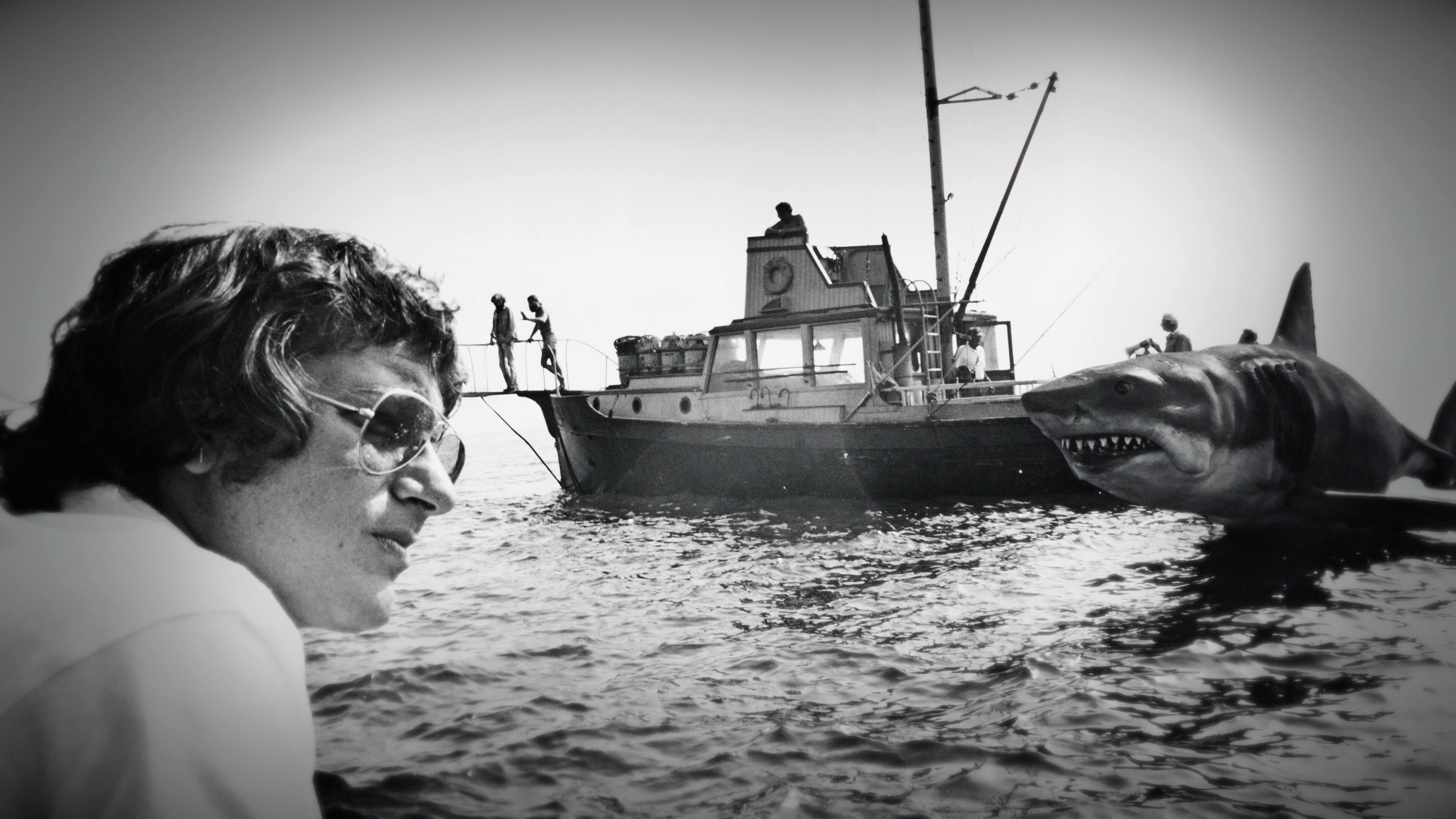 A black-and-white photo of Steven Spielberg, a man in his late twenties with wavy hair, a light skin tone and sunglasses, on a movie set on the water. In the background on the water is a fishing boat and a large mechanical shark.