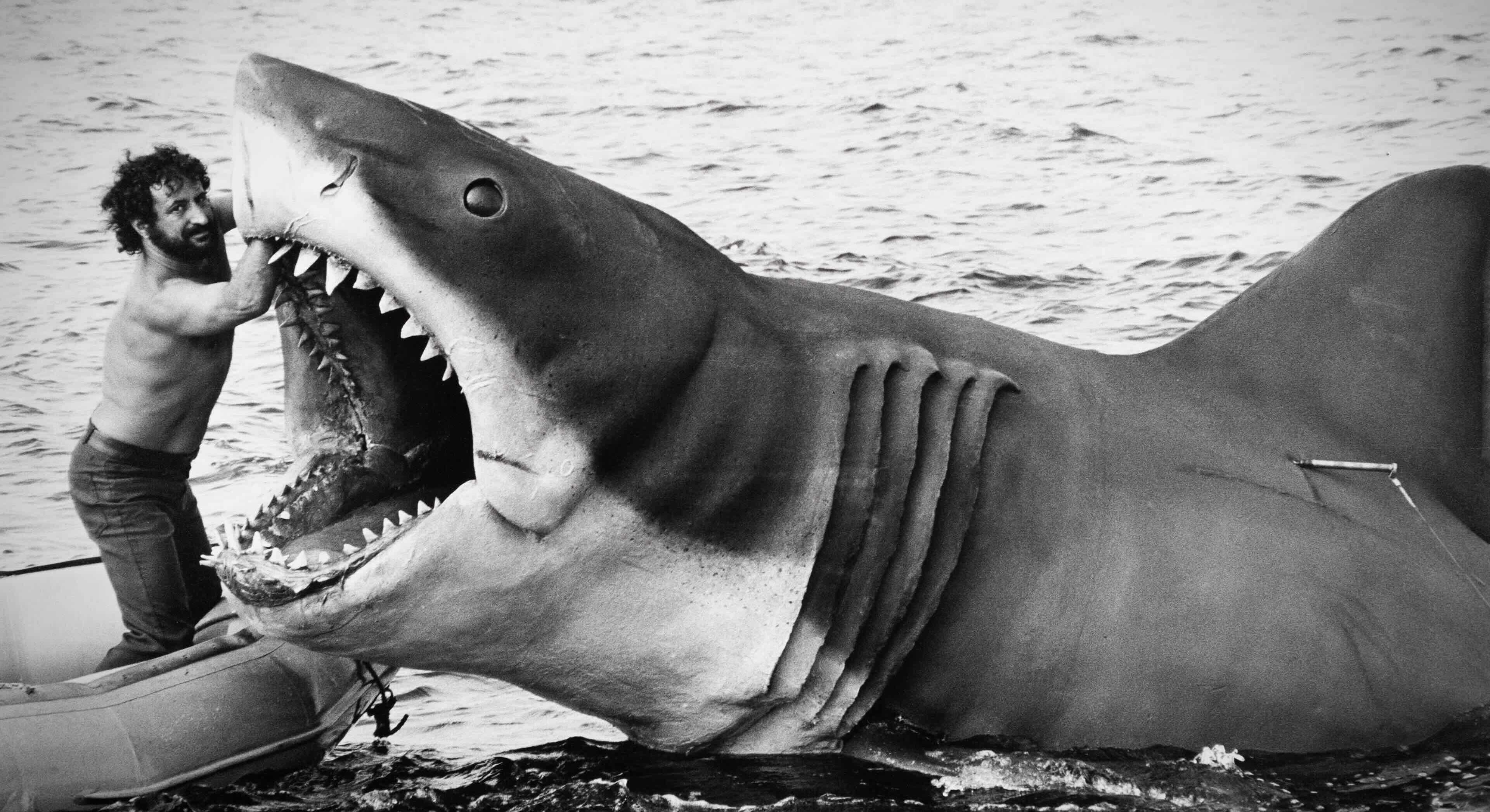 A black and white photo of a shirtless man in jeans holding the open mouth of a large animatronic shark on a movie set near the water.