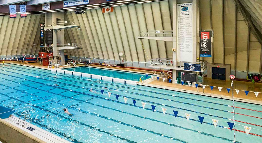 The interior of the Vancouver Aquatic Centre features a 50-metre pool, a beige seating area to the left and a high, pitched roof through which natural light is shining.