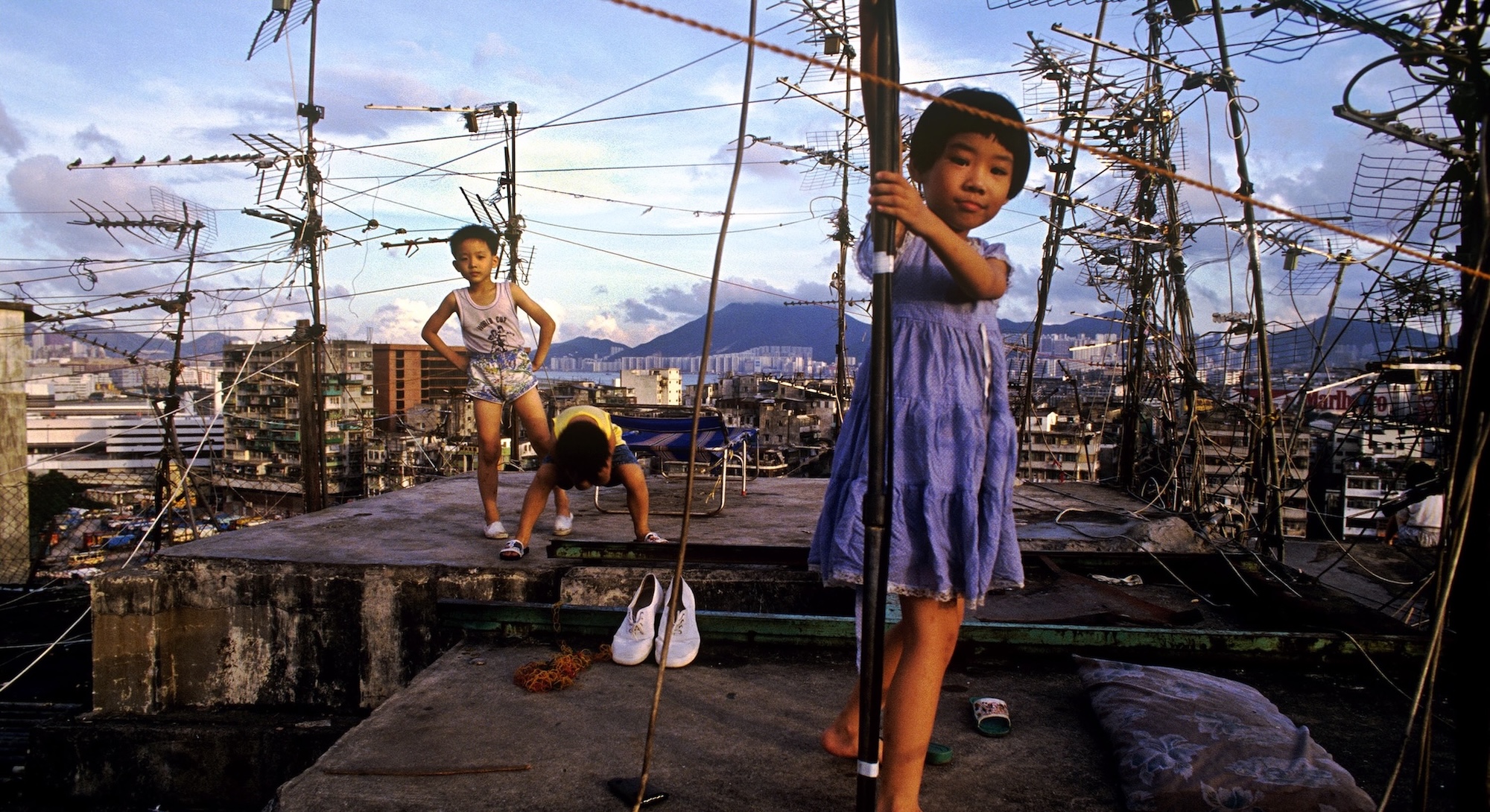 Three young children are standing on the flat concrete rooftop of an apartment building in a dense urban environment. In the foreground is a girl with short dark hair, a medium skin tone and a purple dress. Behind her are two boys in summer clothes; one stands with his hands on his hips, while the other is crouching, looking down.
