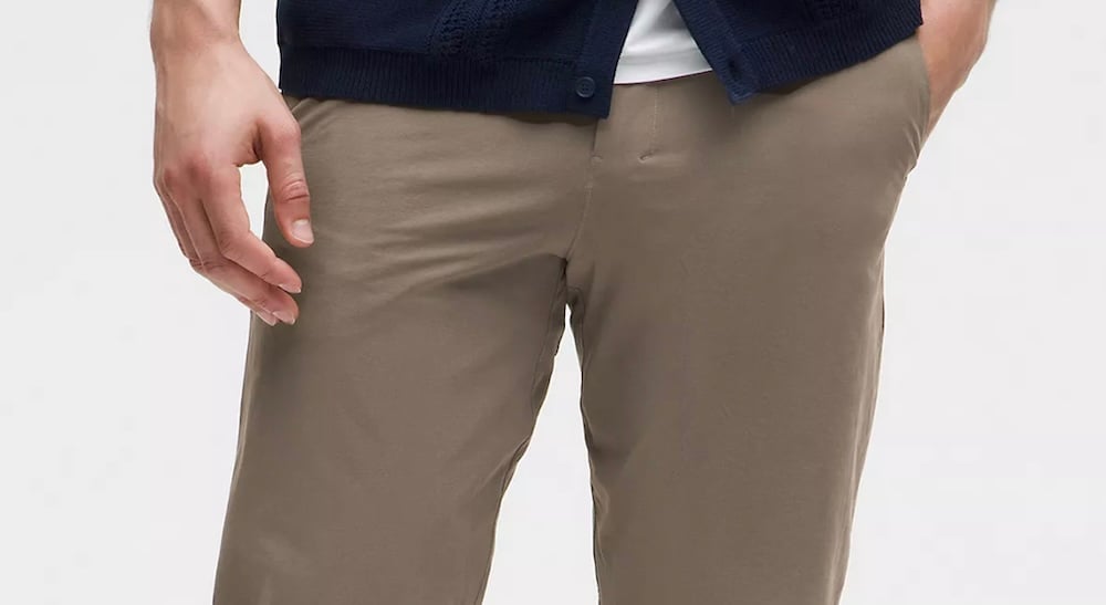 A close-up shot of the waist of a model wearing twill stretch cotton trousers. His hands are resting at his sides and in one pocket. He is wearing a white T-shirt under a dark button-down shirt against a white studio background.