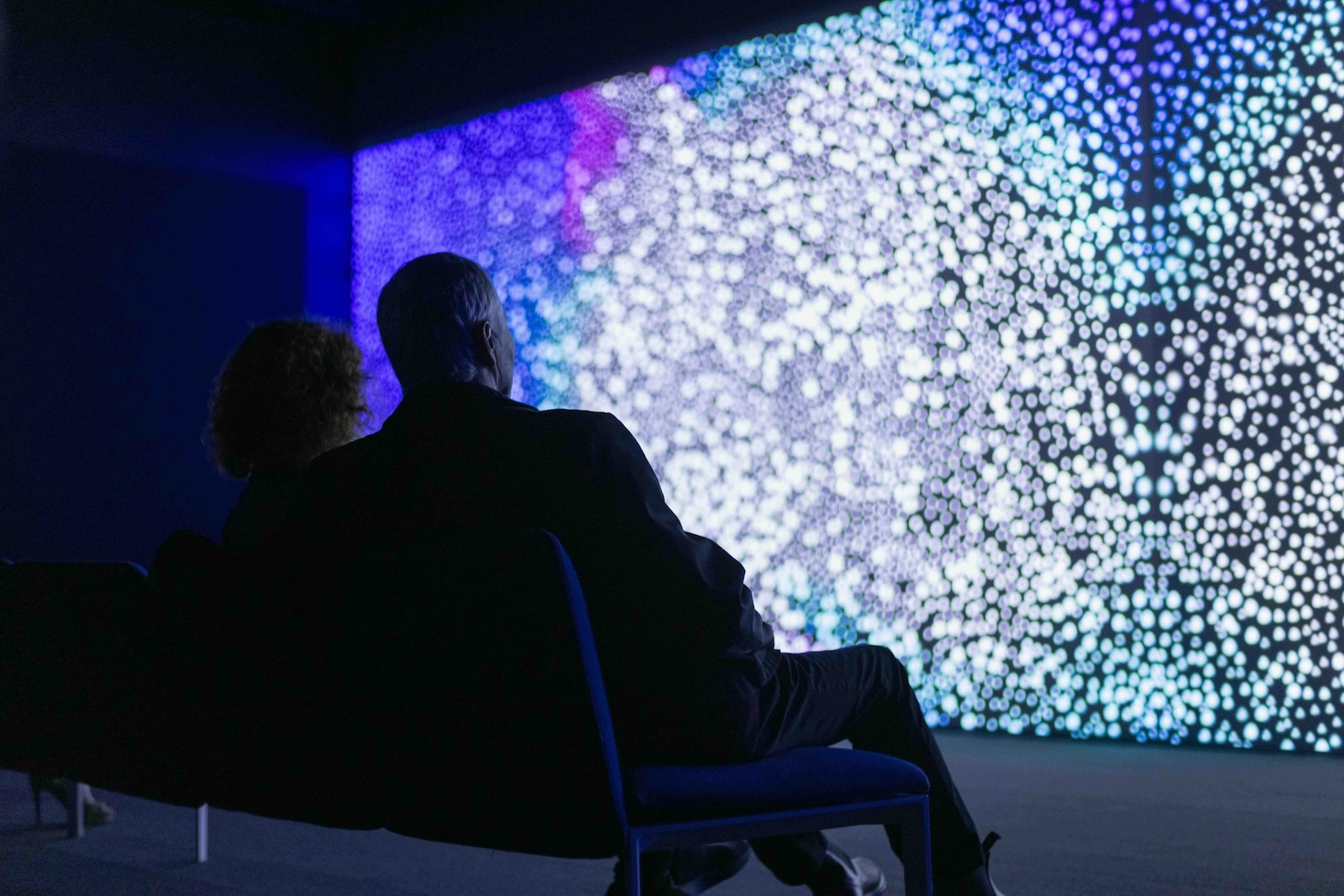 People are seated on a long low bench in a darkened room. In front of them is a wide horizontal screen featuring a digital projection resembling stars in the galaxy.
