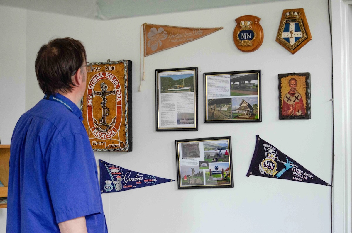A man wearing a blue shirt stands with his back to the camera, looking at a wall displaying flags, framed clippings and mementos.