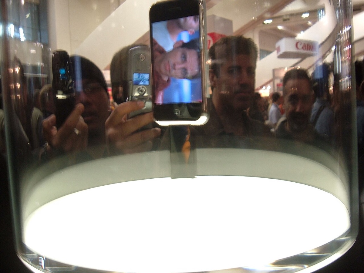 A first-generation iPhone is suspended in a cylindrical glass case. It’s playing a video featuring the actor Ben Stiller. Behind it, a man is taking a photo of the iPhone using a flip phone.