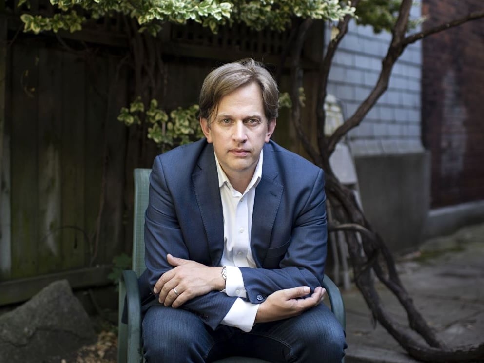 Stephen Marche sits on a chair outdoors with his arms folded on his thighs. He is a light-skinned man with dirty-blond hair, wearing jeans and a blue blazer over a white button-down shirt.