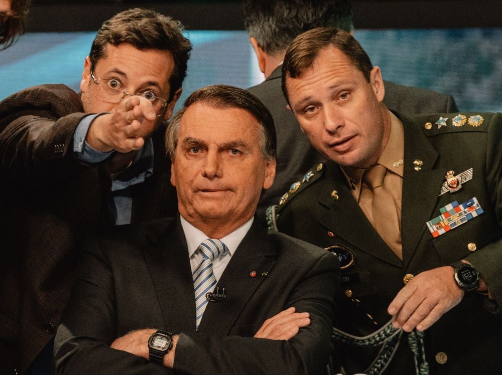 Brazil presidential candidate Jair Bolsanaro crosses his arms as two other political figures lean in toward him, the one on his right passionately pointing at something offscreen.