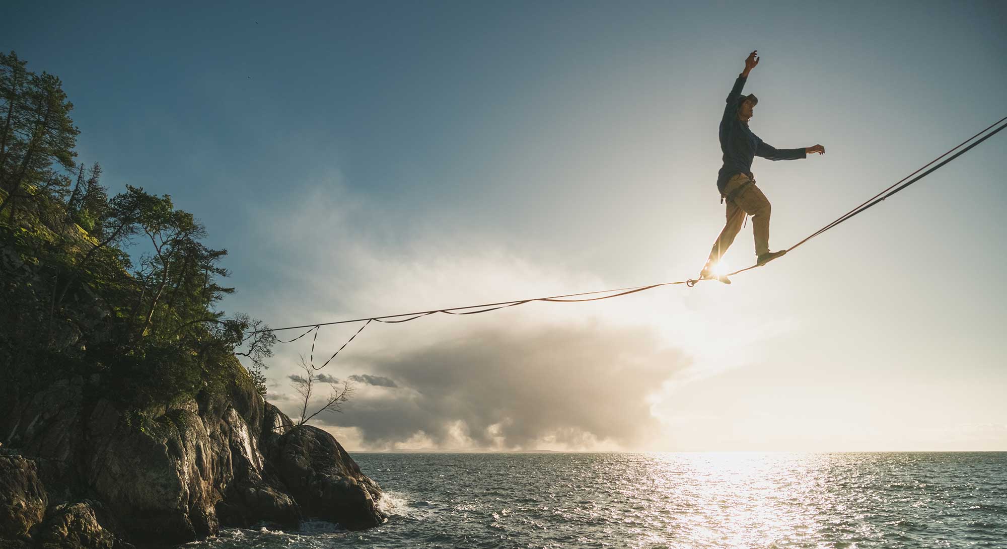 A wide shot of a person walking on a highline over a sunny cliffside ocean. 