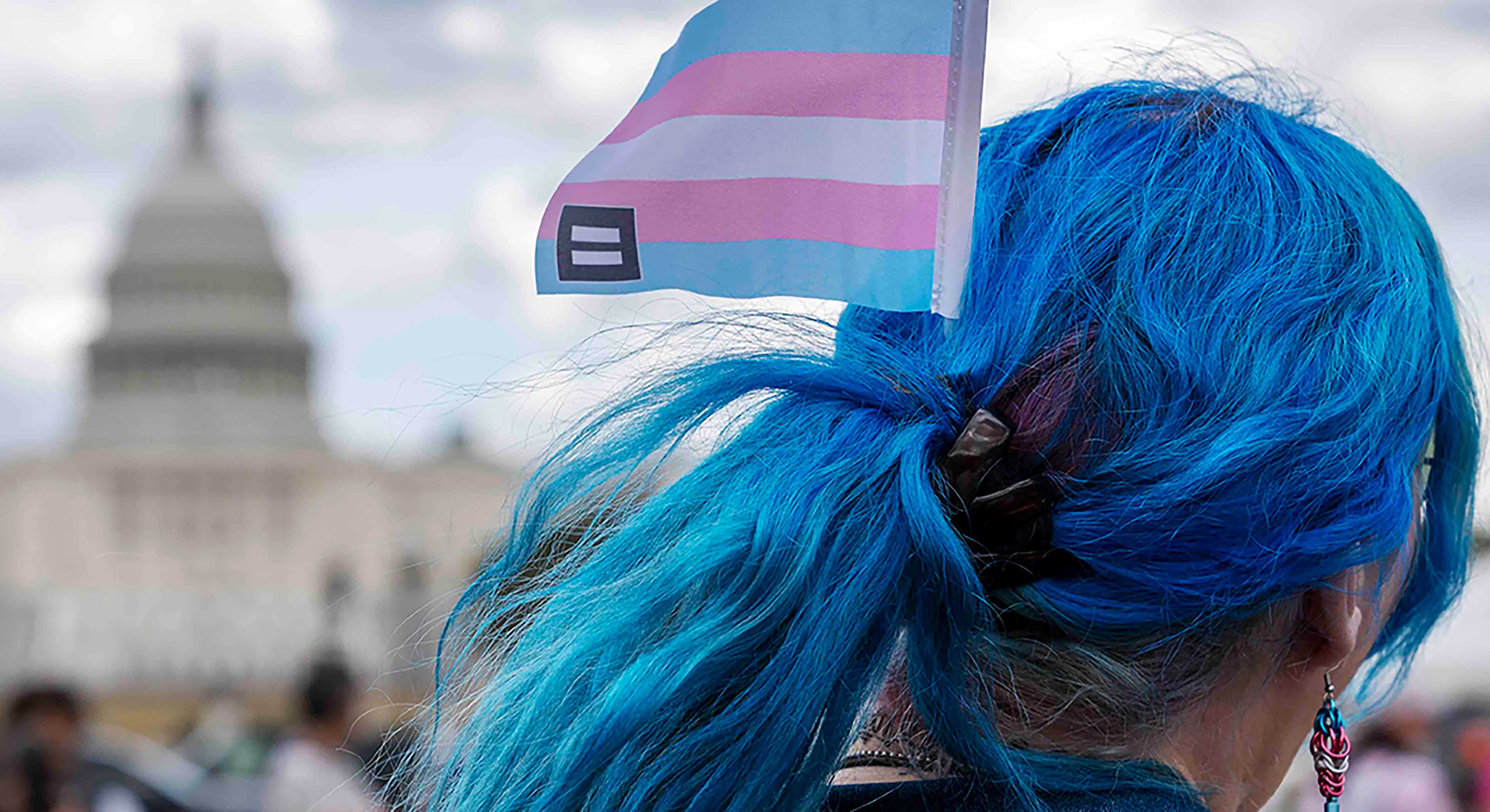 Kali has long wavy blue hair tied back. They are wearing a denim shirt and a dangly earring in blue, pink and white. They are wearing a small transgender flag in their hair. The Washington Capitol is in the background.