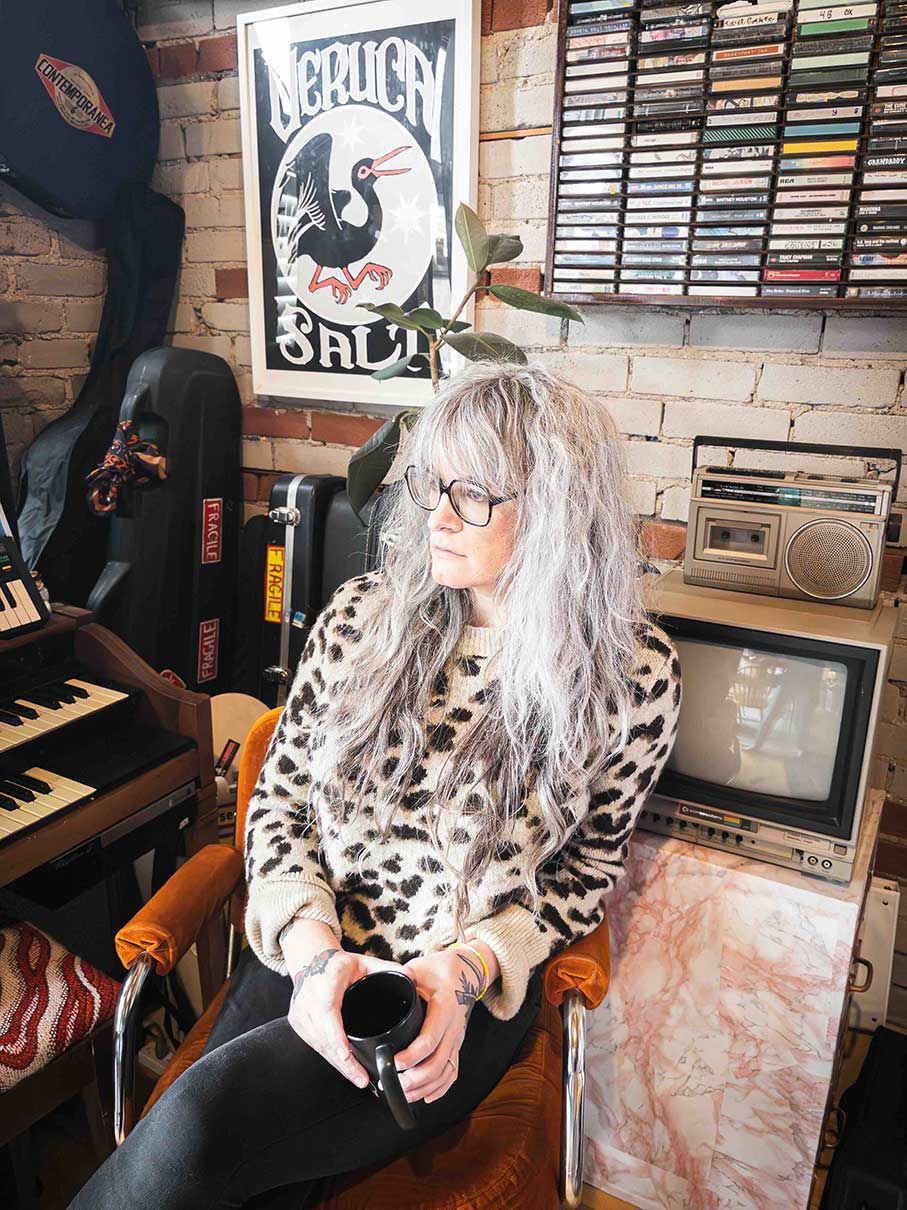 Niko Stratis has long wavy hair dyed light lavender. She is seated in an indoor space in front of a small vintage television and beside a keyboard. Behind her is a shelf of cassettes. She is wearing a leopard-print sweater and holding a black coffee cup.