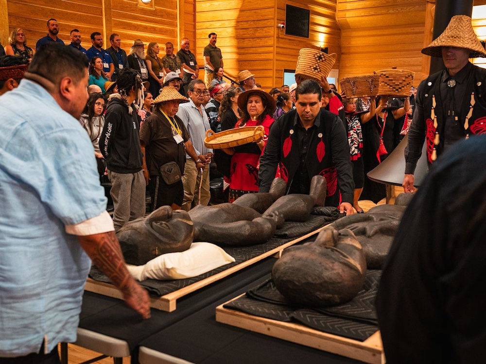 People are standing in an indoor space with wooden walls. Some people are wearing cedar hats and ceremonial First Nations regalia, or holding cedar baskets aloft. In the foreground a group of men arranges two carved wooden figures on a wide table.