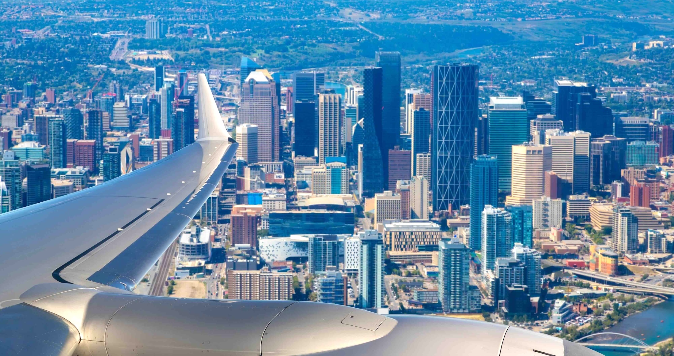 Highrise towers in downtown Calgary span out against a blue sky. In the foreground is the silver wing of a passenger jet.