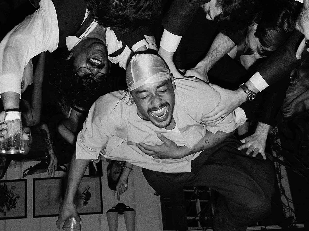 A black-and-white photo depicts an upside-down shot of Dijon Duenas crowd-surfing atop a crowd of people in formal evening wear. Duenas has a dark skin done and a moustache. He is wearing a light headscarf and T-shirt, holding a beverage glass and laughing.