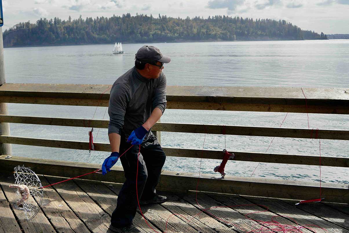 George Ying is wearing a grey long-sleeved athletic shirt, a grey cap and sunglasses. He is wearing blue work gloves and in the midst of casting a crab trap off a wooden pier and into the water in front of him.