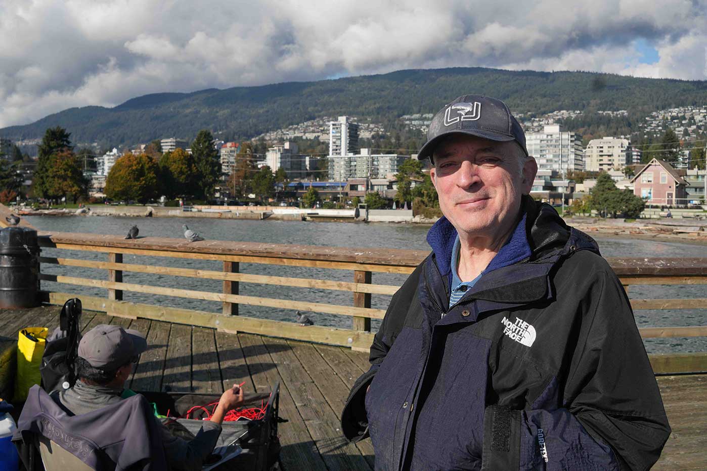 Tony Kondaks stands on a wooden pier with water and an urban setting behind him. He is wearing a ball cap and a navy jacket. His hands are in his pockets and he smiles at the camera. Another crab fisher is seated behind him.