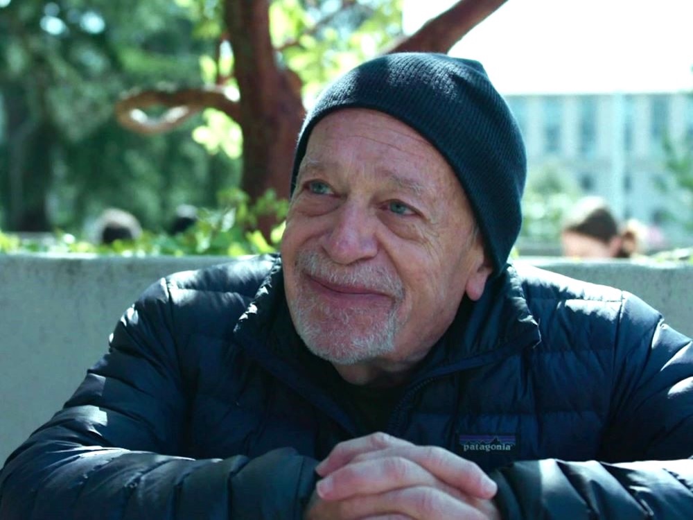Robert Reich, a man in his 70s with light skin tone, blue eyes and a close-cropped grey beard, looks toward the left of the frame. He is wearing a navy blue toque and Patagonia puffer jacket.