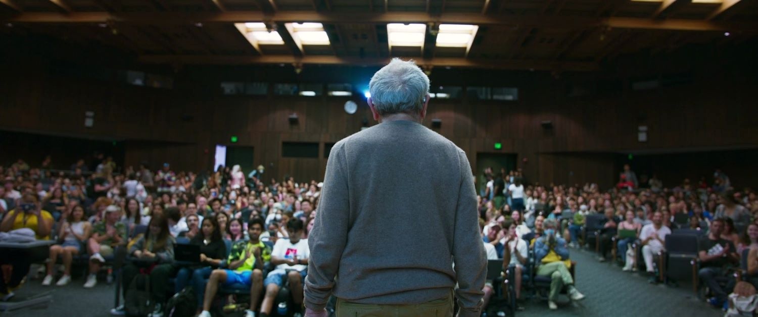 A man with short grey hair wearing a grey sweater is seen from behind, standing before a large lecture hall of people.