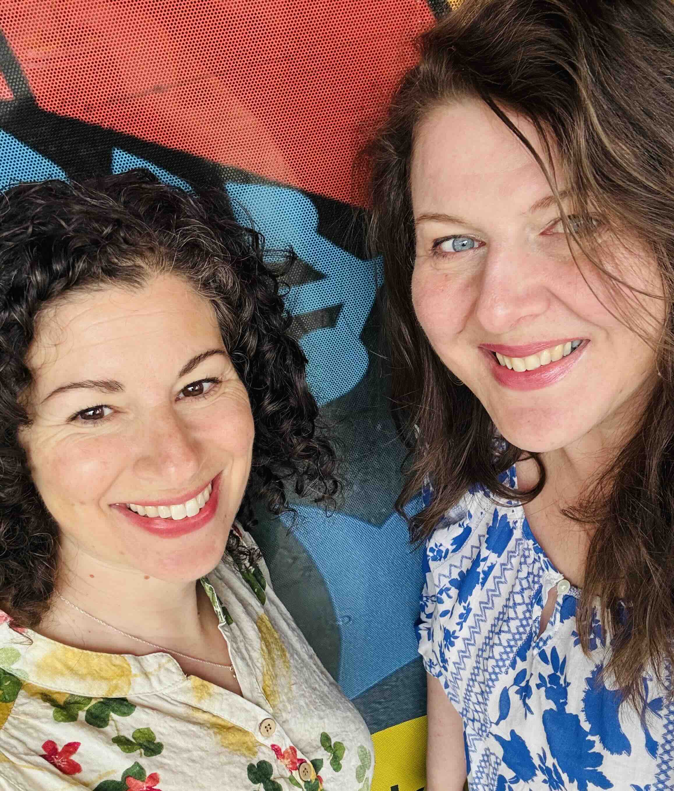 Claire Sicherman, left, has dark curly hair and is wearing a light floral too. Nicole Breit, right, has shoulder-length brown hair and is wearing a blue and white patterned top. They are smiling at the camera and standing against a colourful mural background.