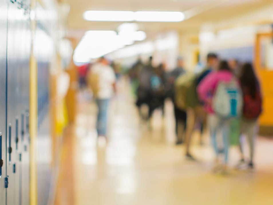 A soft-focus photograph of a high school hallways depicts students gathered in the middle of a corridor with blue lockers on the left side of the frame.