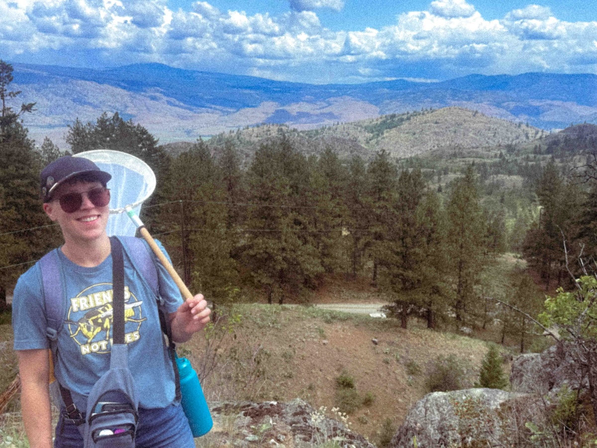 Jade Lee has short hair and is wearing a navy ball cap, sunglasses and a blue T-shirt. She is holding a white net with a wooden handle over her shoulder and she has a water bottle and fanny pack slung around her body. She stands outdoors in a wooded area; behind her is a dry mountain range and a clear sky with fluffy clouds.