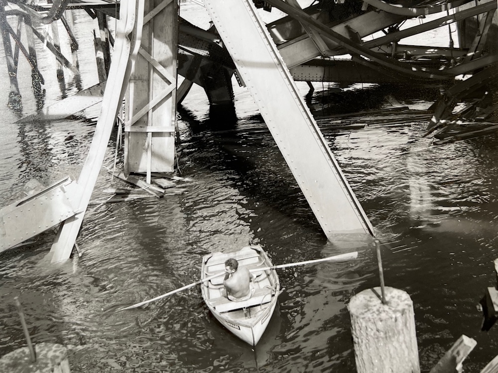 A sepia-toned black and white photo of a shirtless man in a small rowboat surrounded by twisted metal beams.
