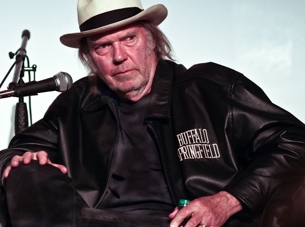 Neil Young, a scraggly dark-haired late-middle-aged man wearing a straw hat and a dark jacket that says &lsquo;Buffalo Springfield.&rsquo;