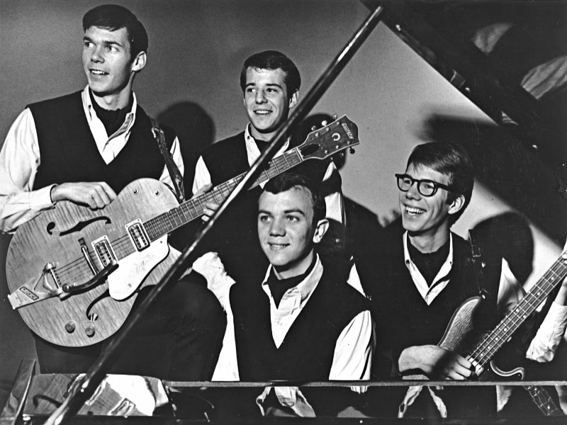 A black and white publicity photo of a rock and roll foursome of teenaged white males holding their instruments and smiling.