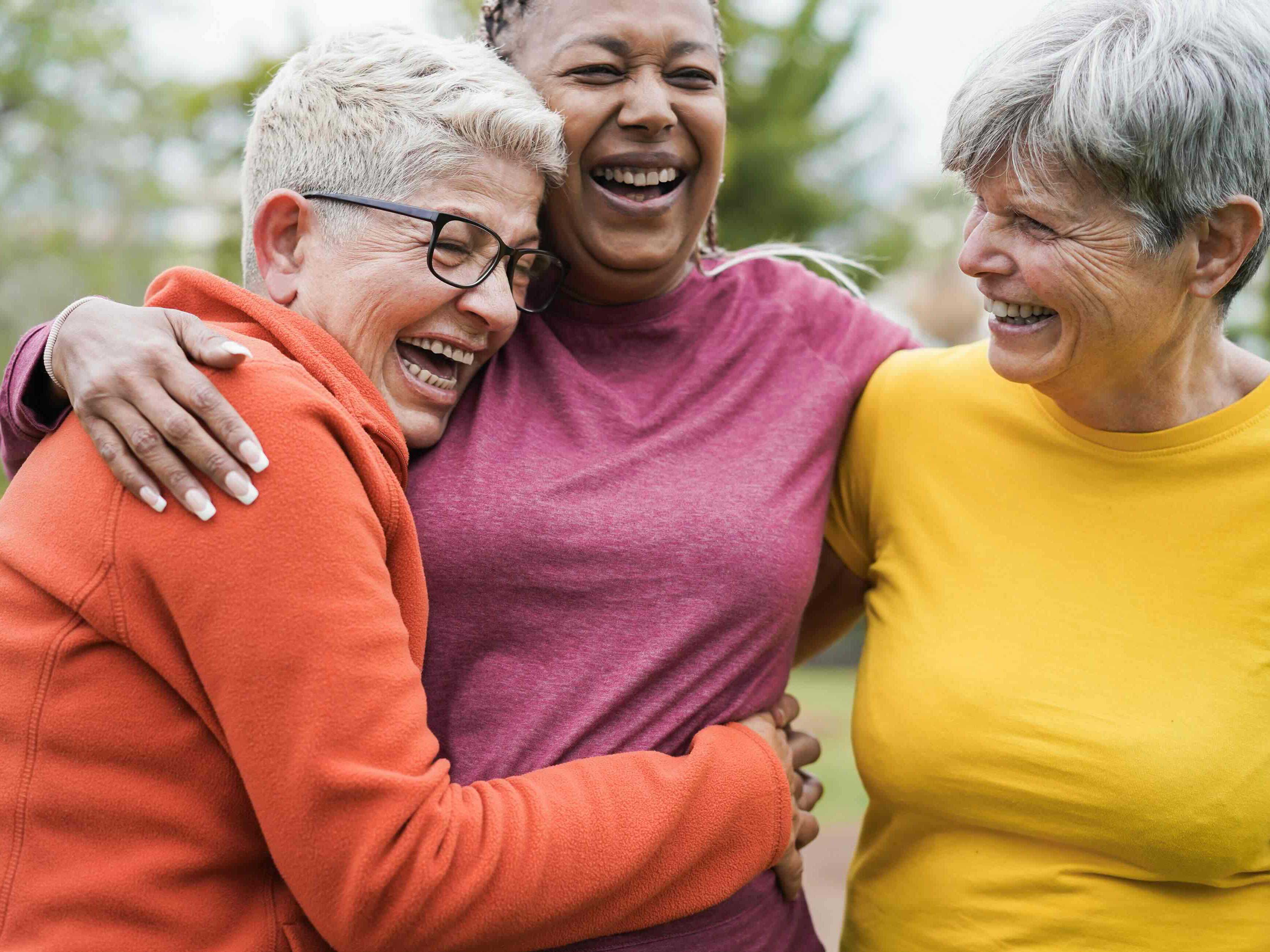 Three older women embrace each other while laughing and smiling. They are standing outdoors. From left to right, they are wearing orange, fuschia and yellow. 