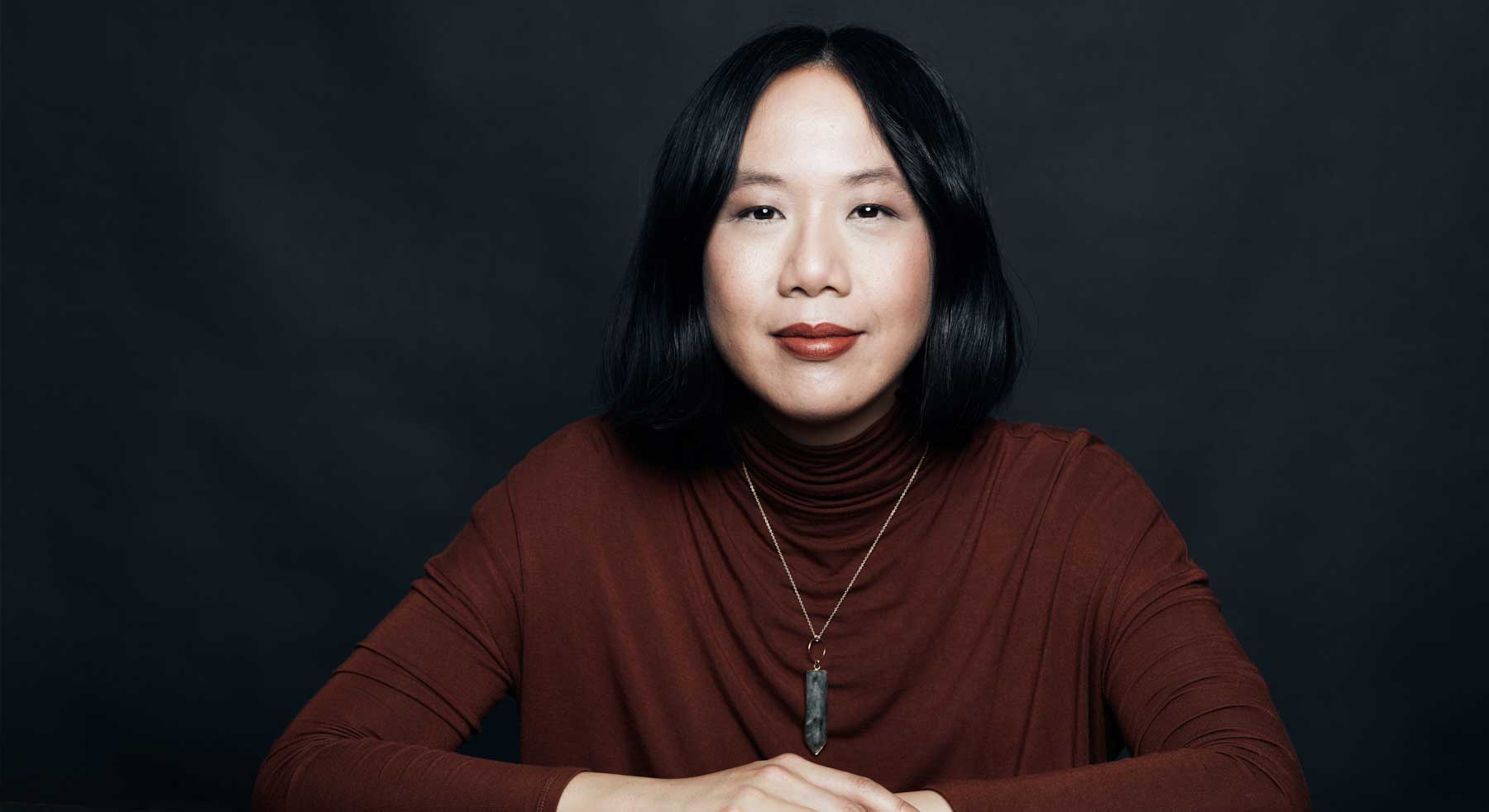 Jen Sookfong Lee is seated against a dark studio background with her hands folded across the table in front of her. She has a pale skin tone and black shoulder-length hair. She is wearing a burgundy sweater that matches her lipstick. She is wearing a necklace hung with a grey semi-precious stone.