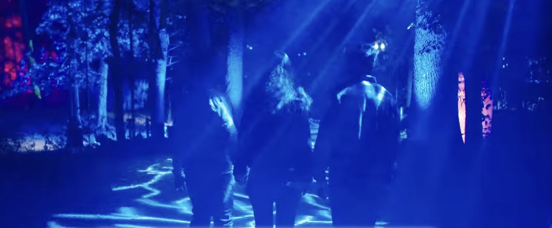 Three people walk through a dark, blue-lit forest.