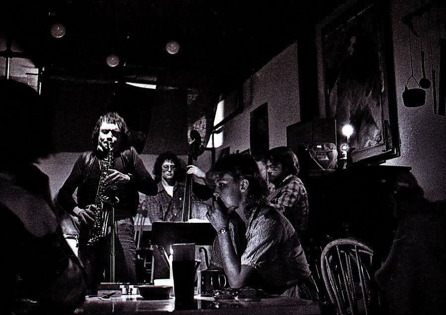 A dark and grainy black and white photograph shows a man playing the saxophone. Behind him someone plays stand-up bass and people sit at tables in a bar-like atmosphere.