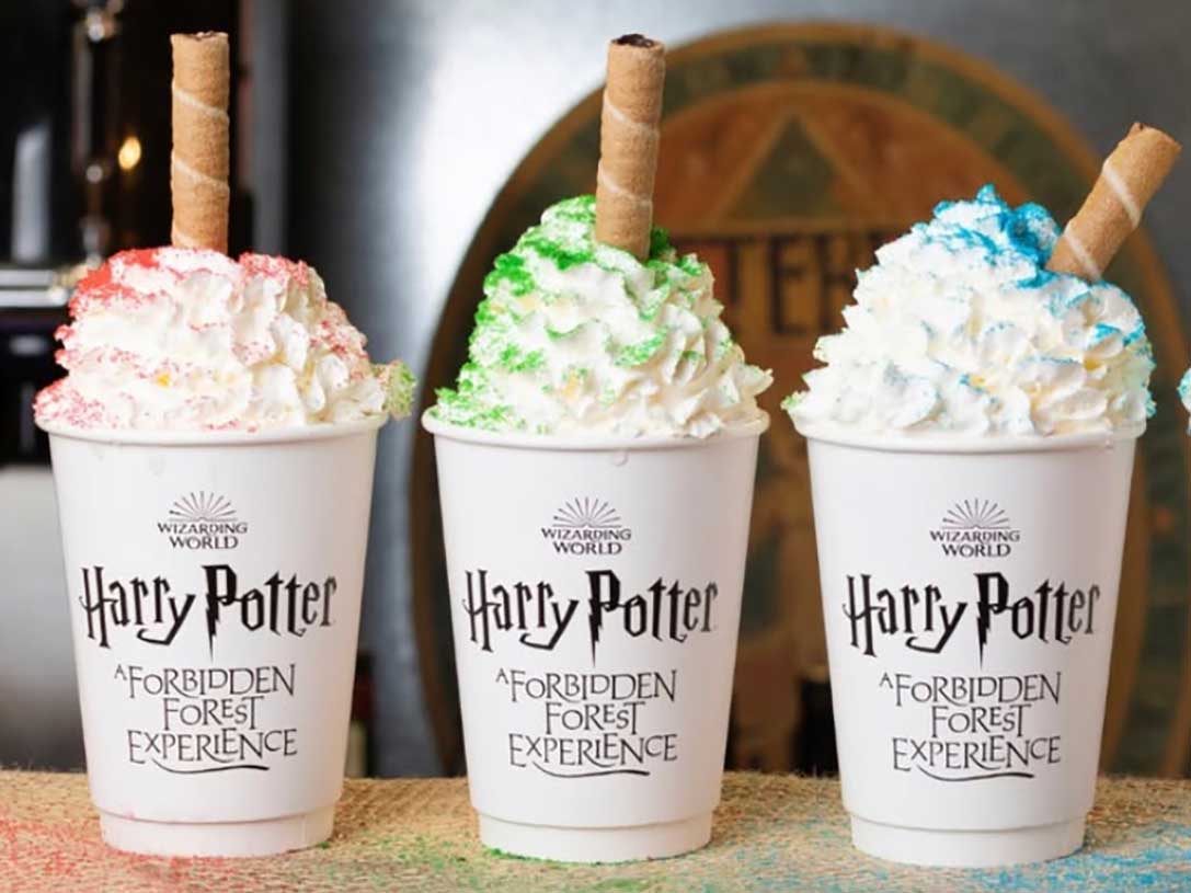 Four cups of hot chocolate are lined up on a counter. Each is topped with whipped cream and a tube cookie. Each has been topped with a different colour of sugar to represent the four different Hogwarts houses.