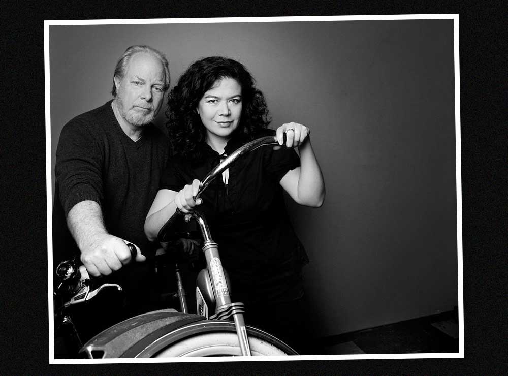 A black and white image of an older man with white hair and beard standing behind a woman with dark hair who is holding the handlebars of an old-fashioned bicycle.