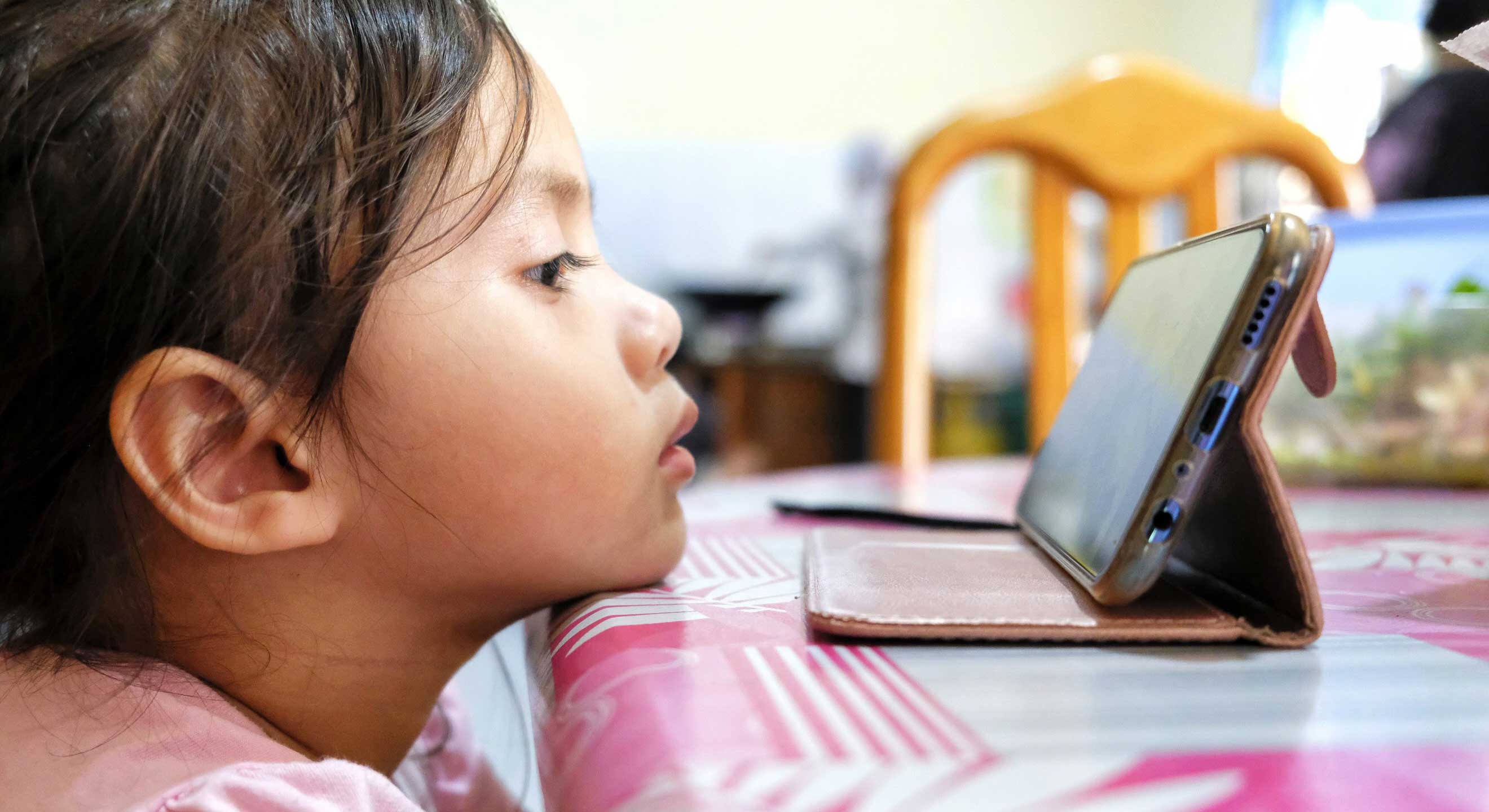 A young girl to the left of the frame rests her chin on a tabletop covered with a pink and white vinyl covering. She is looking at a smartphone perched towards her against its carrying case. She has dark hair, a medium-skin tone and is wearing a pink shirt. In the background is a plastic container of food on the table and an adult, out of focus, to the right.