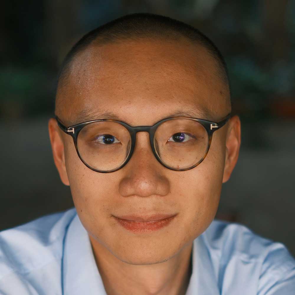 Dan Wang has short black hair and tortoiseshell glasses. He is wearing a light blue button-down shirt and is seated against a grey studio background.