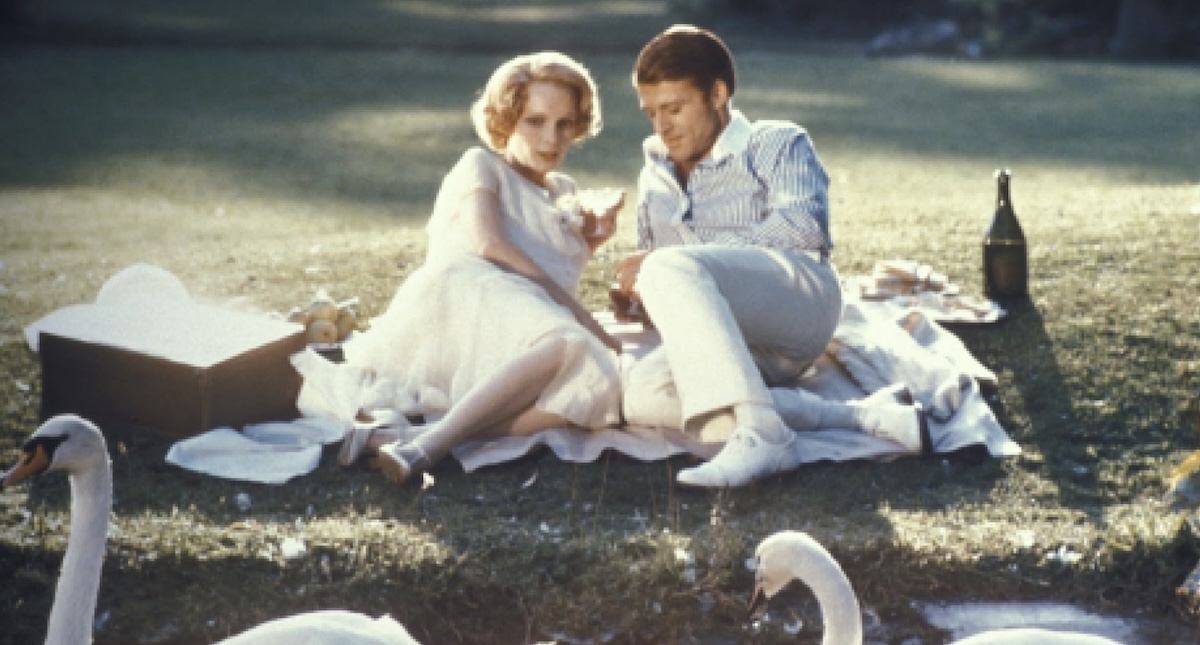 Two actors lie facing each other on a white picnic blanket in the grass with swans in the foreground. Mia Farrow, left, is wearing a white dress. Robert Redford, right, is wearing a blue-and-white-striped shirt and white trousers.