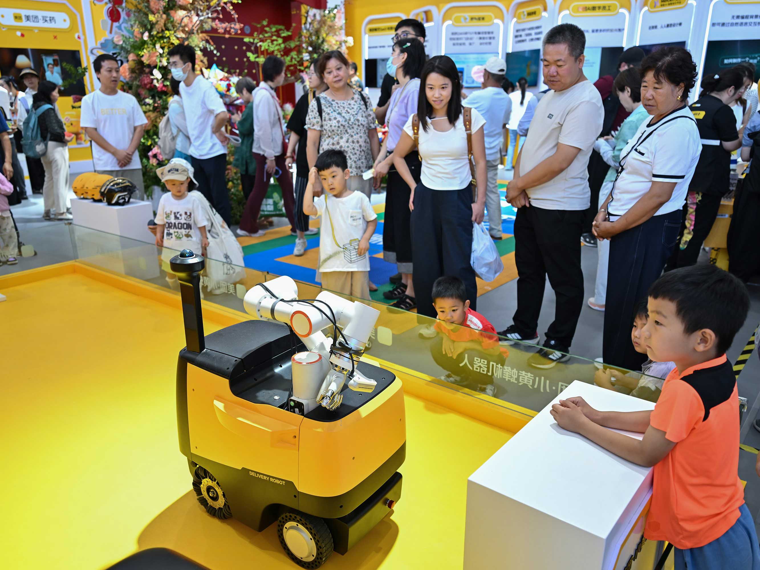 People in white T-shirts and casual summer apparel gather around a demonstration of a yellow robot, a cart-like structure, carrying white cylindrical shapes across a yellow floor. Surrounding the front of the display are several young children.