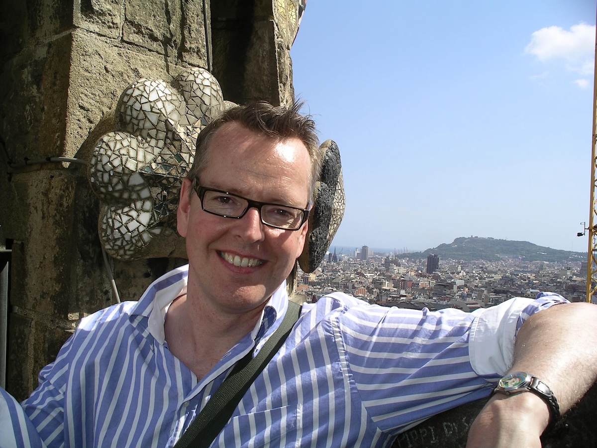 Steve Burgess has short spiky brown hair and glasses. He&rsquo;s smiling and wearing a blue-and-white-striped button-down shirt. He is leaning against a concrete ledge and a sprawling city is visible in the background under a blue sky.