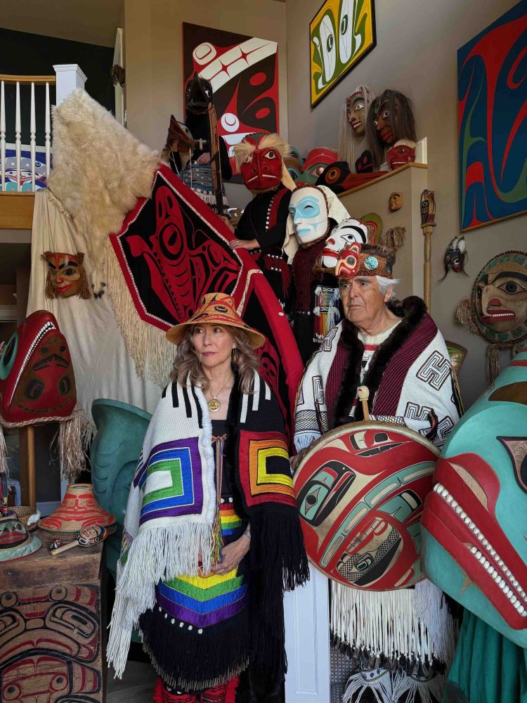 A photograph depicts two people in Haida regalia among ceremonial masks, drums, blankets and artwork in an indoor space. Terry Lynn Williams, left, and Robert Davidson, right, stand at the bottom of a staircase and three people in masks stand behind them on the stairs.