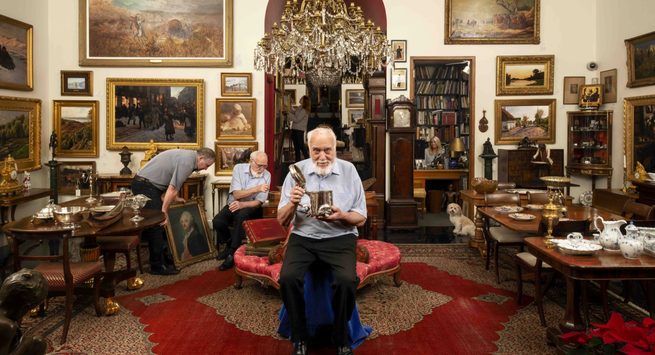 An abstract photograph depicts an elderly man seated on a stool covered in royal blue fabric in the centre of the frame. He has white hair and a white beard, and he is wearing a short-sleeved light blue shirt and black trousers. He is seated under an ornate crystal chandelier in a room full of mahogany furniture and pieces of art on the wall in gold frames. He is holding a silver vessel with the lid open. Behind him are other adults interacting with artwork in the room.