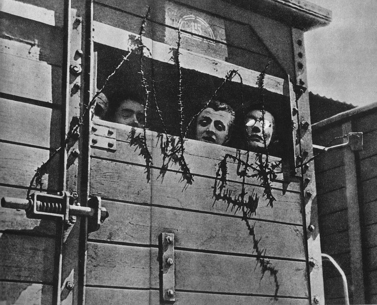 An old black and white image of three people looking out of a small opening in a wooden railcar strung with barbed wire.