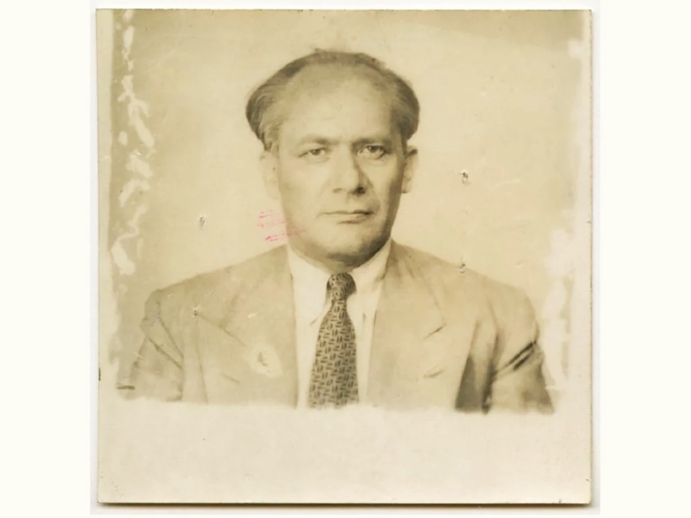 An old, blemished, sepia-toned image of a man with thinning hair wearing a suit and tie.